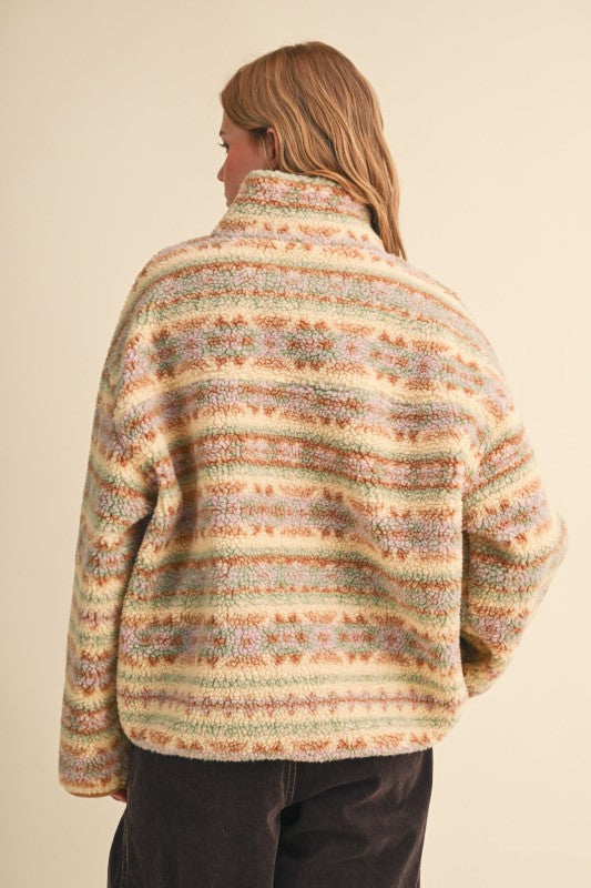 A woman shown from the back wearing a fuzzy knit mock-neck sweater with horizontal multicolor stripes in beige, green, orange, and brown tones, paired with dark pants against a plain cream background.